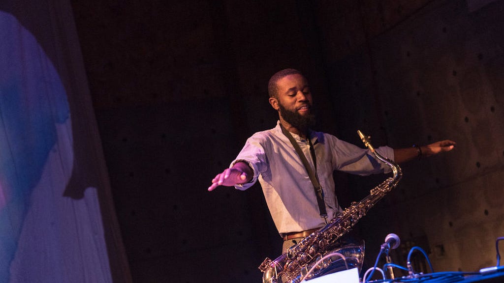 photo of Jerome Ellis, a Black man with very short hair and a long beard. He looks down at his arms outstretched on either side of him and has a saxophone in front of him.