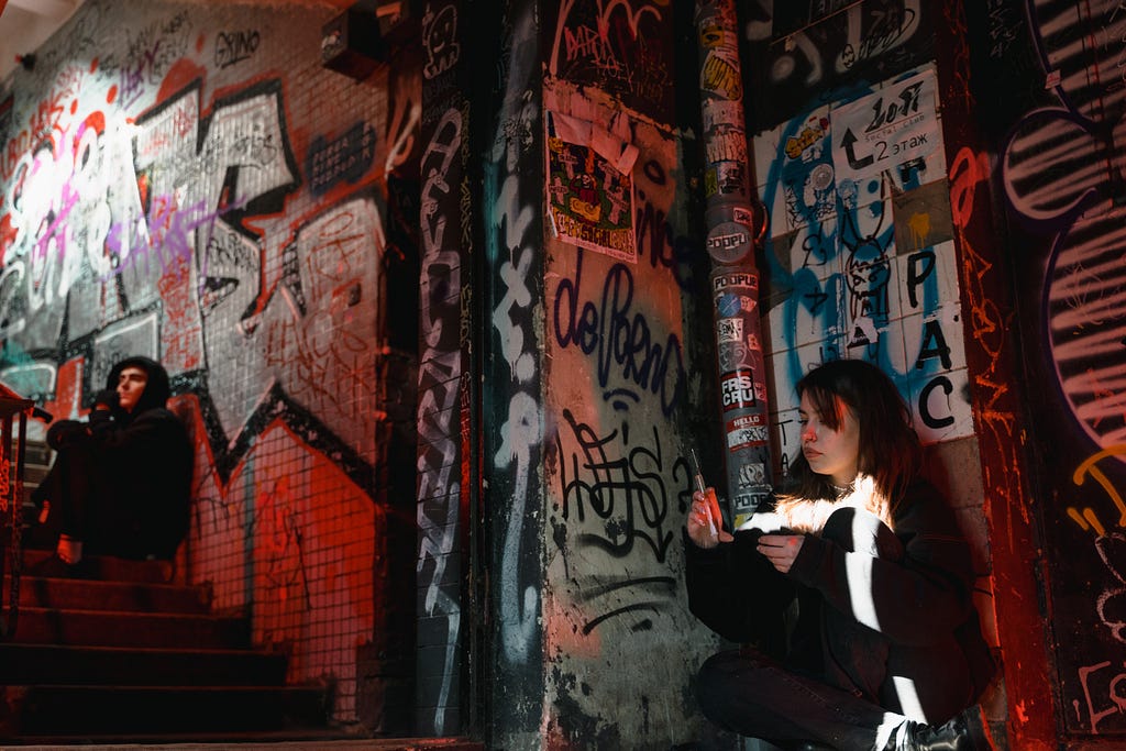 A woman and a man sit inside a subway station taking drugs. They sit against a graffiti-filled wall.