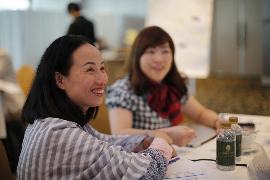 Two women smile as they sit at a conference room table holding bottled water and stacks of paper.