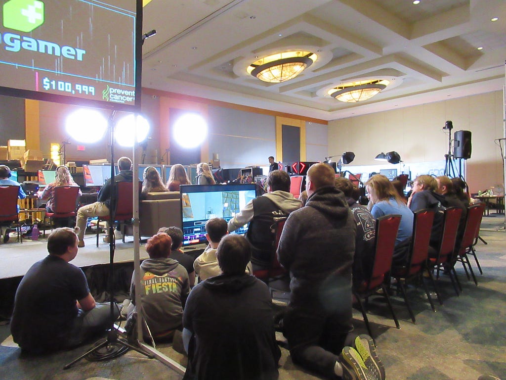 A gathering of people behind the stage of GDQ around the backup couch. The backup couch is positioned in front of a TV.