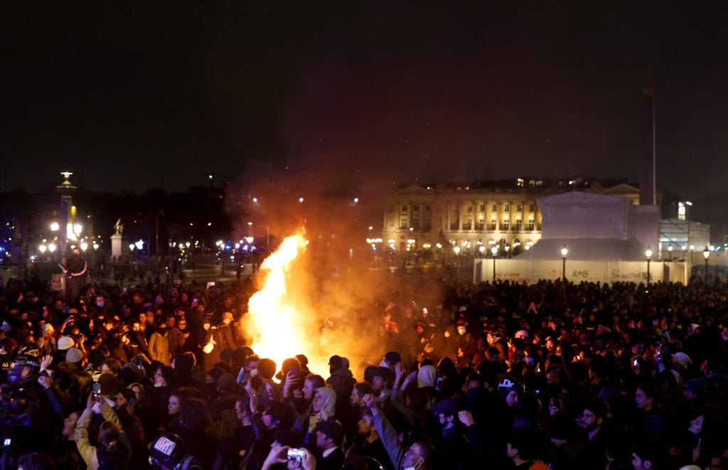 Picture of packed public square in France. It is night, and a fire blazes in the middle of the group.