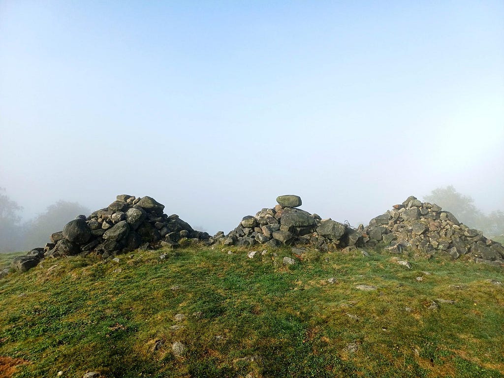 3 stone cairns, close together in a row. The cairns are made of small gritstone boulders piled up into triangular shapes.