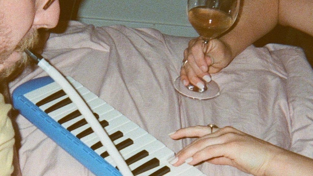 A couple plays a melodica together while drinking wine.