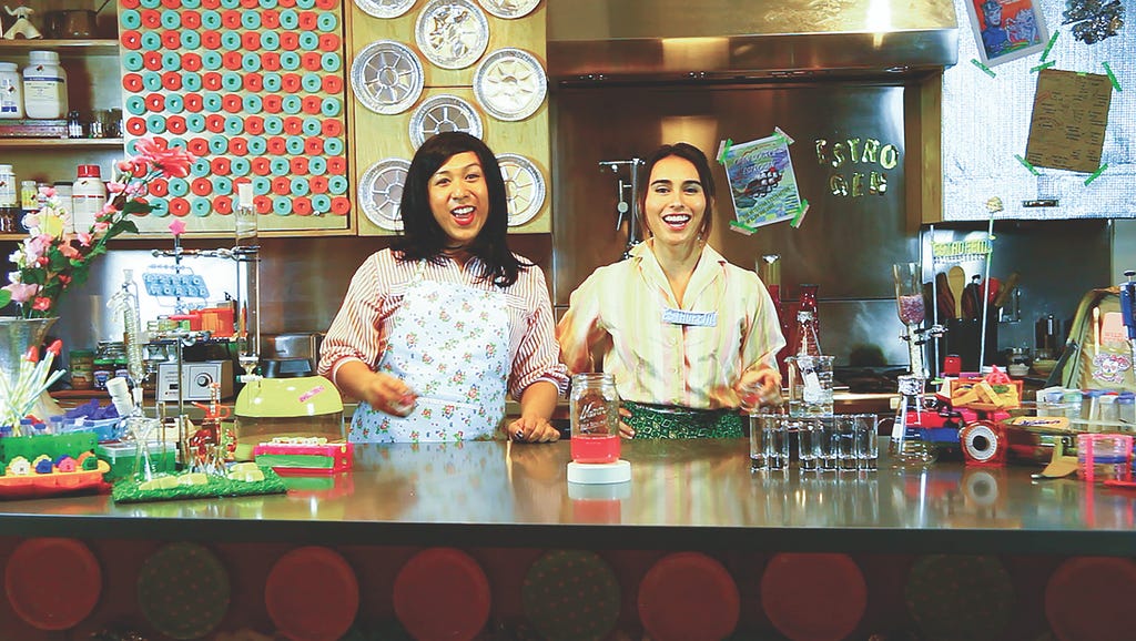 Two television hosts stand in bright and colorful kitchen with science lab kit making homemade estrogen hormones