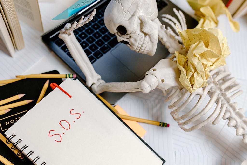 Skeleton figurine with its head on a laptop on a white table and crumpled pieces of paper lying on him
