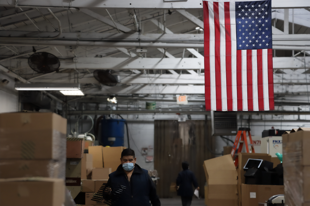 A man walks toward the camera through a warehouse and passes under an American Flag hanging from the rafters of the plastic comb factory. He is wearing a blur surgical mask as a woman walks away from him and the camera.