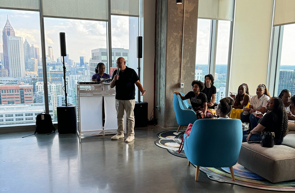 Google VP welcomes guests to their downtown office during a meet & greet event with attendees sitting in chairs listening to a. DJ.