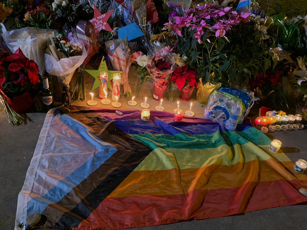 LGBTQ inclusive flag on the ground surrounded by candles and flowers.