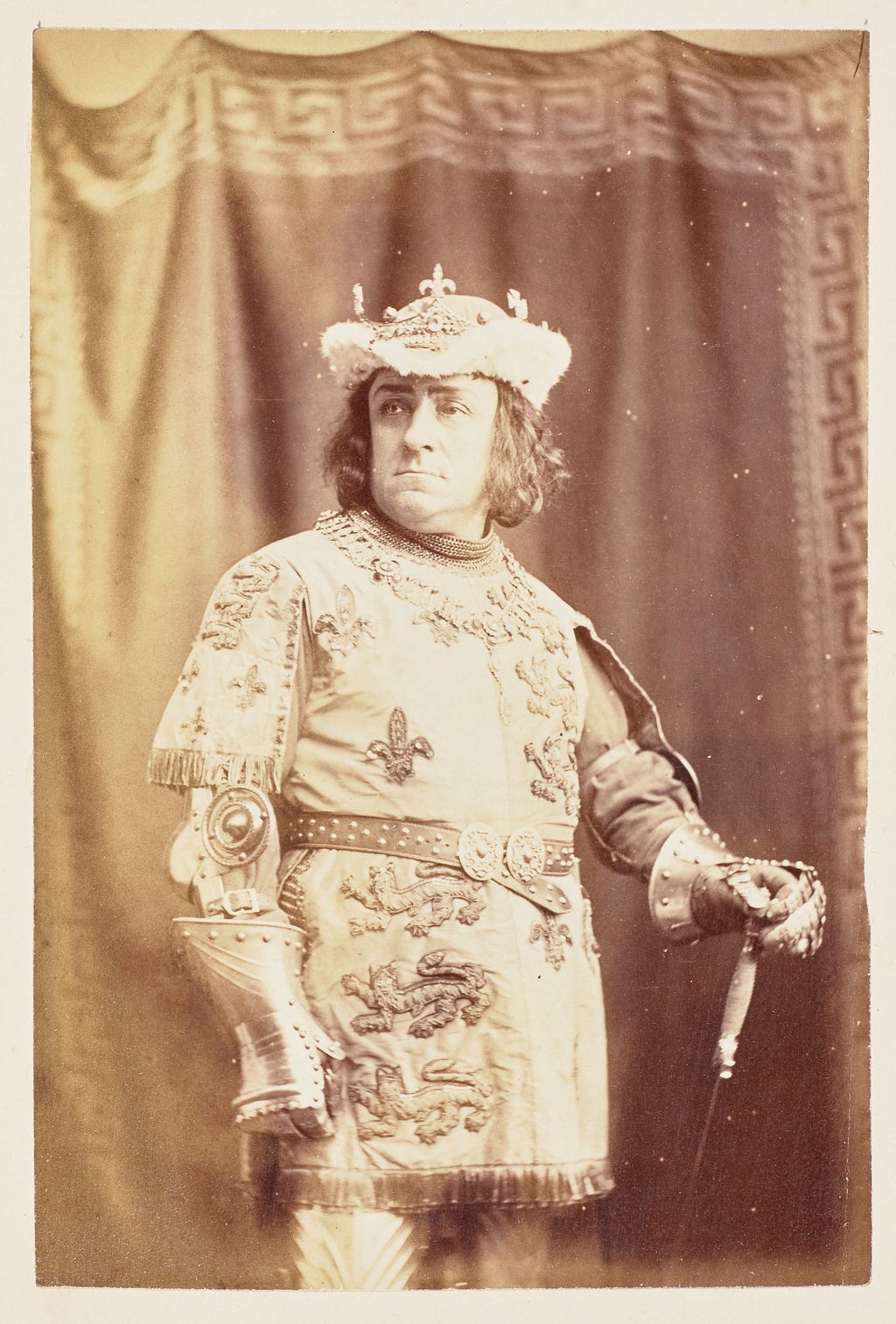 Black and white photo. Man in rich 15th-century dress. Belted tunic decorated with fleur de lys and lions, hat and gauntlets.