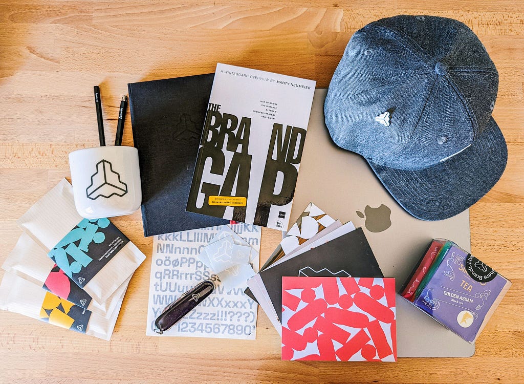 An assortment of Frontify branded onboarding item, including a cap, chocolate, notebook, teas, Swiss army knife, and a book called “The Brand Gap.”