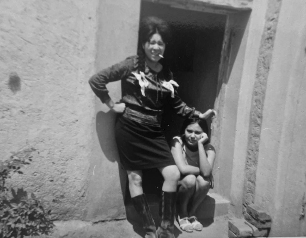Black and white photo of two young women in a doorway, one standing with a cigarette in her mouth and the other sitting with her head in her hand, both looking at the camera