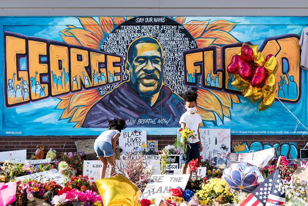 An image of a mural of George Floyd with people setting flowers and signs beneath it.