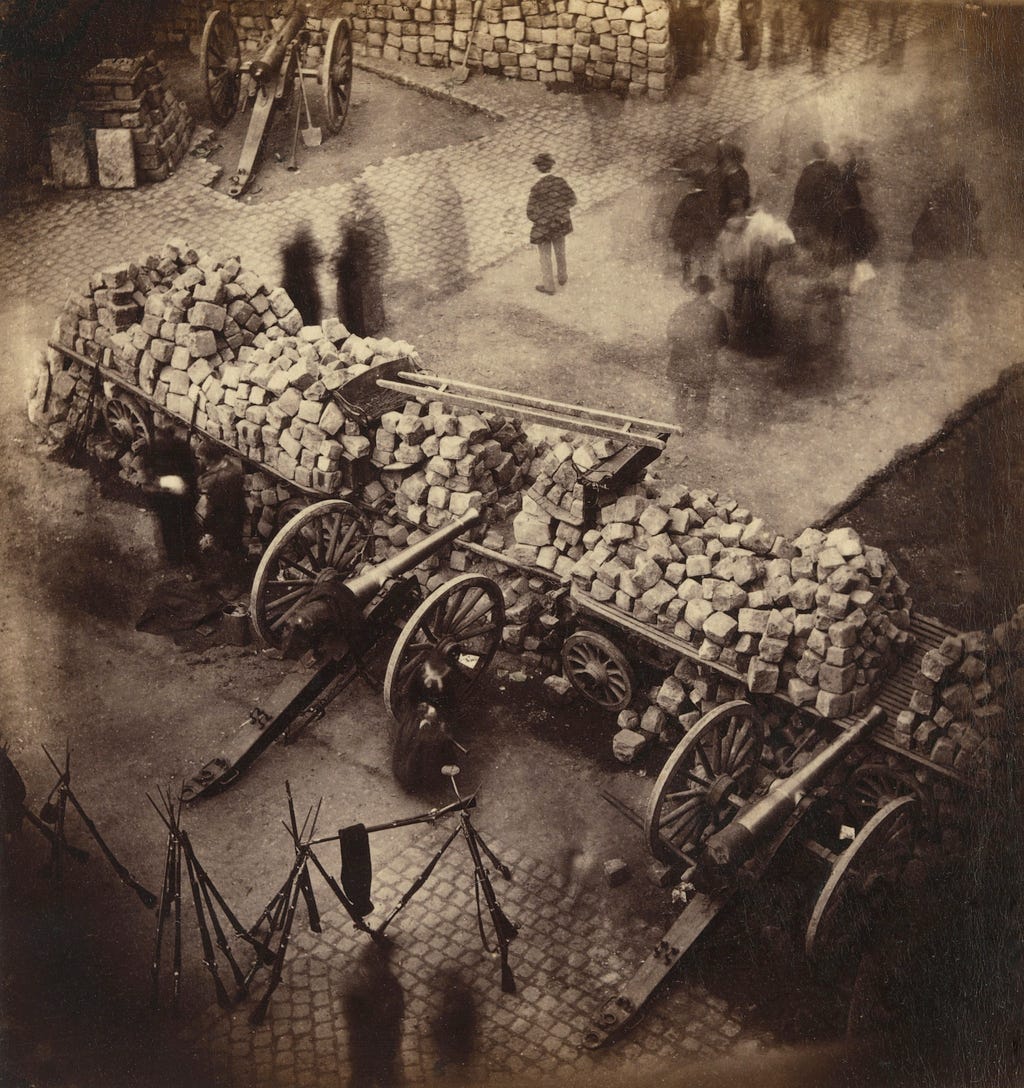 A photograph, taken from a building overlooking a barricade of the Paris Commune, which has been assembled from street paving stones and cannons. Rifles are stacked to the lower left, while in the upper right several figures appear. Because it is a 19th century photograph, with a long exposure time, and the people must have been moving around as the photograph was taken, they appear as ghostly figures.