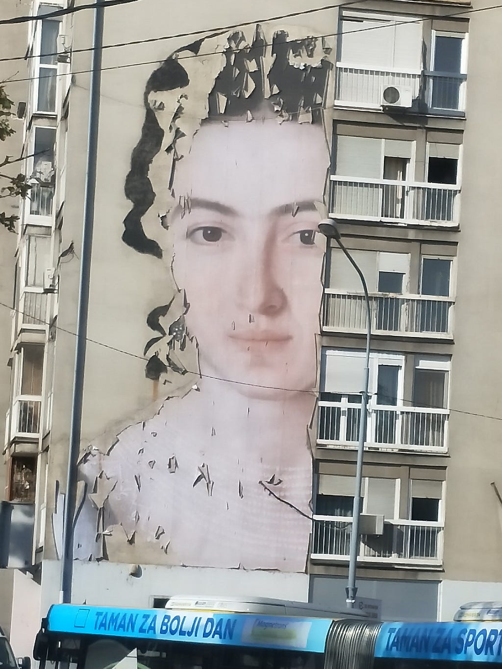 Street art, a simply painted woman’s face gazes serenely from the wall of a dull apartment block, edged with peeling paint.
