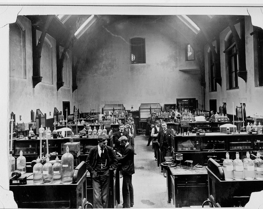 Black and white photograph of Owen’s College Chemistry Laboratory. Several students can be seen standing in a large hall with tall ceilings. There are several desks topped with large glass jars and scientific equipment.