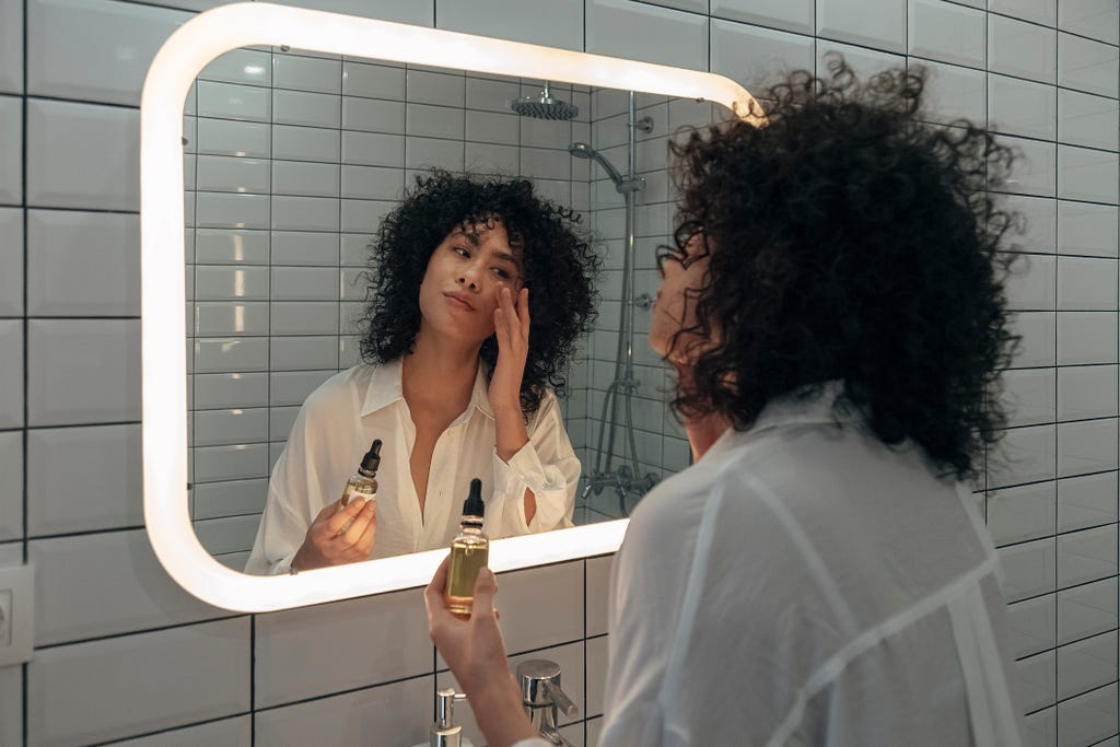 Woman in a light cotton shirt and curly dark hair applies skin serum in a subway-tiled bathroom mirror