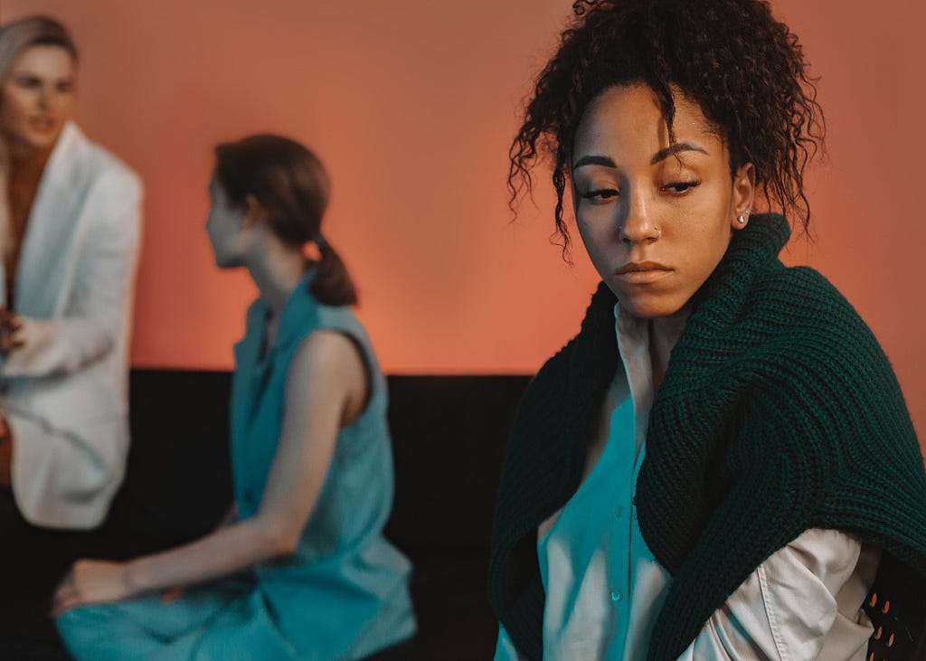 Black girl sitting alone with two white women talking in the background