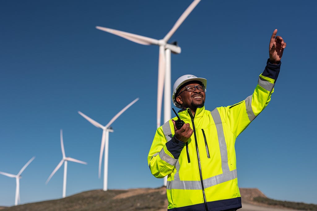 With four windmills behind him, a man in a hardhat and bright yellow safety jacket holds a walkie-talkie while pointing towards the sky.