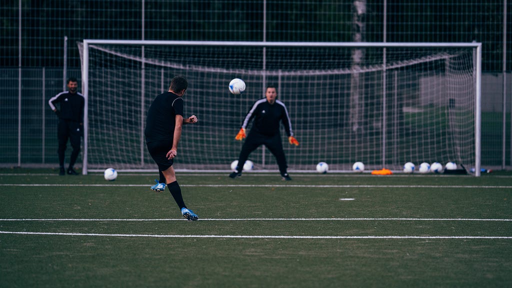 A soccer player in all black practicing shooting on a goalkeeper with orange gloves in goal.