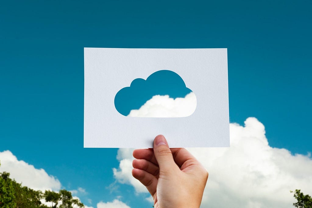 A hand holding a piece of paper with a cloud cut on it, against a cloudy, blue sky.