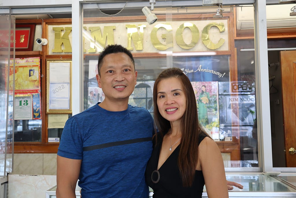 Phillip and Lynette stand side by side facing the camera and smiling in front of a glass case and a large gold sign that reads “Kim Ngoc” in all caps. Phillip wears a blue t-shirt, and Lynette wears a black sleeveless blouse. Lynette has long, straight, amber colored hair and is about 5 inches shorter than Phillip, who has short, straight, black hair.