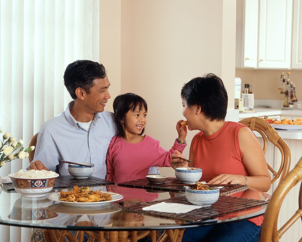 An Asian family, an adult male and female are seated around a table eating a meal with a young female standing in between the