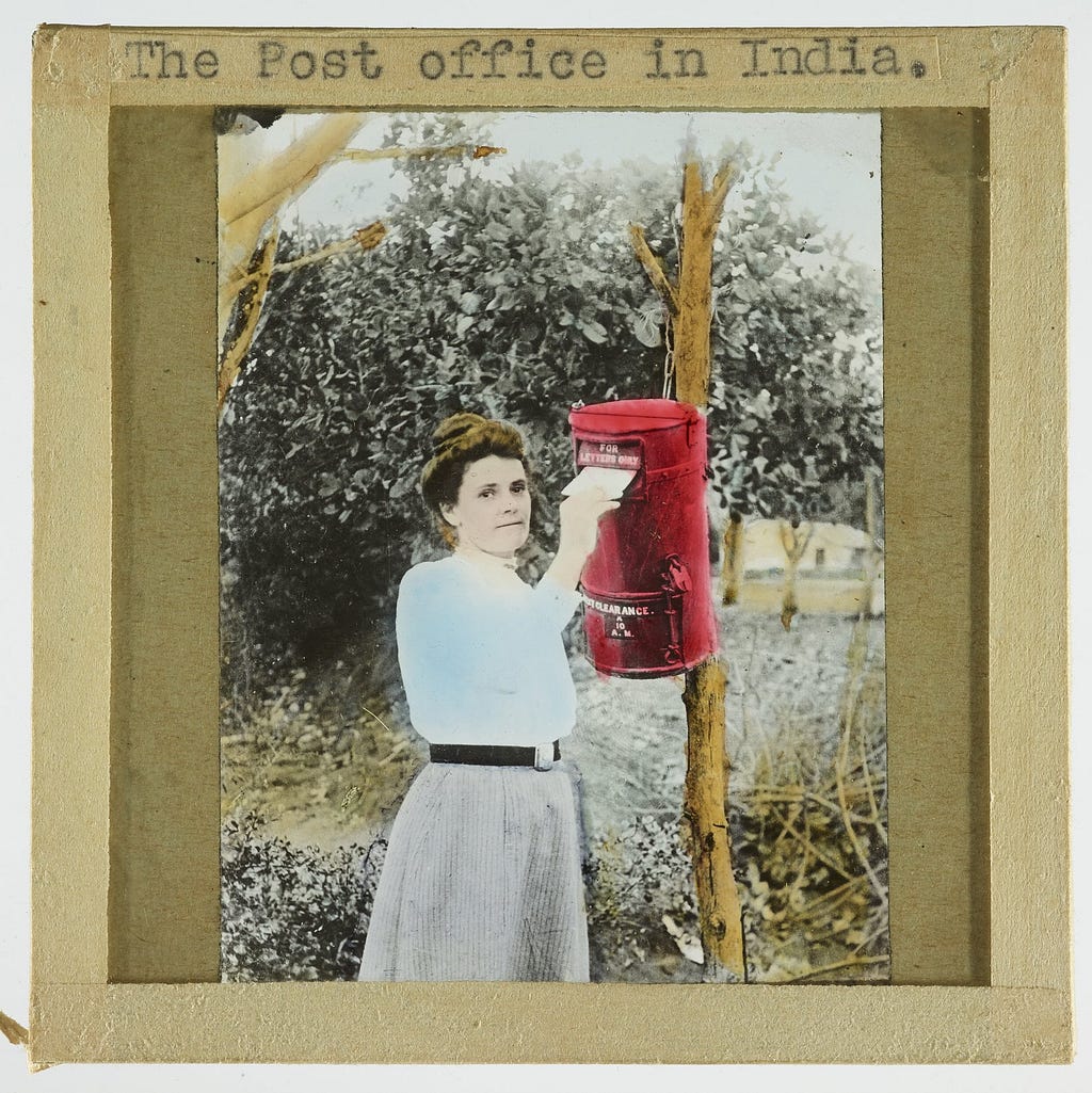 A woman in Edwardian dress posts a letter into a red postbox.