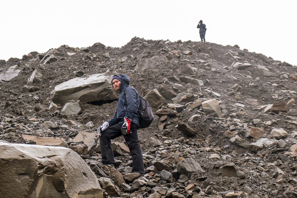 Sean Auffinger climbs rocky terrain in Svalbard, and turns towards the camera at the base of a hill. Another person stands at the top of the hill in the distance.