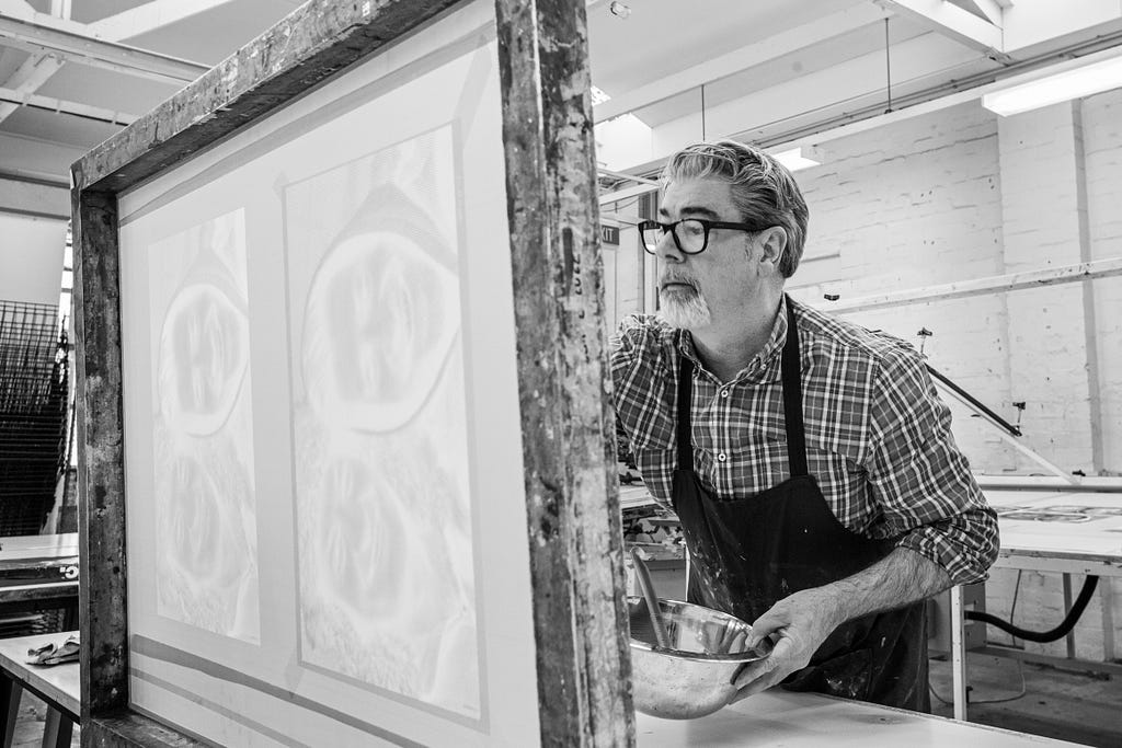 A black and white photograph of Richard Harding working on a new artwork in the printmaking studio of RMIT. He is wearing a checkered shirt and apron and has a large wooden screen in front of him.