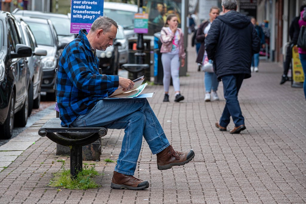 Rick, a middle aged white man, wearing a blue and black checked shirt and blue jeans, sitting on a black bench looking through an activity pack. People and signs on the High Street behind him.