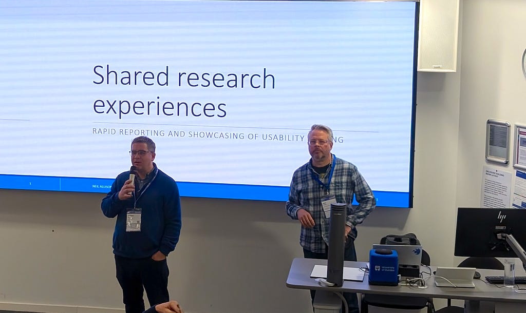 Organiser, Andrew Millar, introducing presenter, Neil Allison. Both are standing in front of a screen with the presentation title ‘Shared research experiences’,