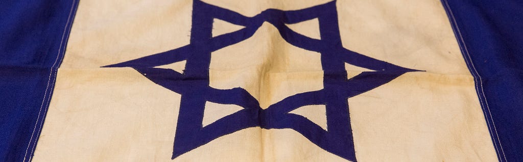 A hand-sewn flag, feature a blue Star of David at the center and two horizontal blue cloth stripes near the top and bottom.