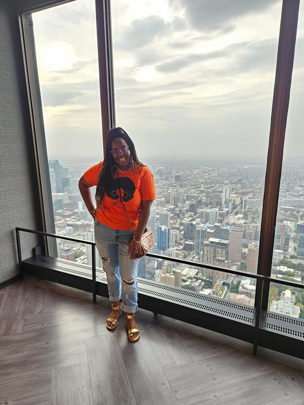 Kara taking a picture in front of the Chicago skyline. She is wearing a orange shirt and ripped blue jeans. Her shirt has an image of a black woman with an afro and hoop earrings. She is wearing gold open-toed sandals with red painted toes. Kara is also wearing a crossbody bag with flowers on it. Kara’s hair is in braids and in a high ponytail.