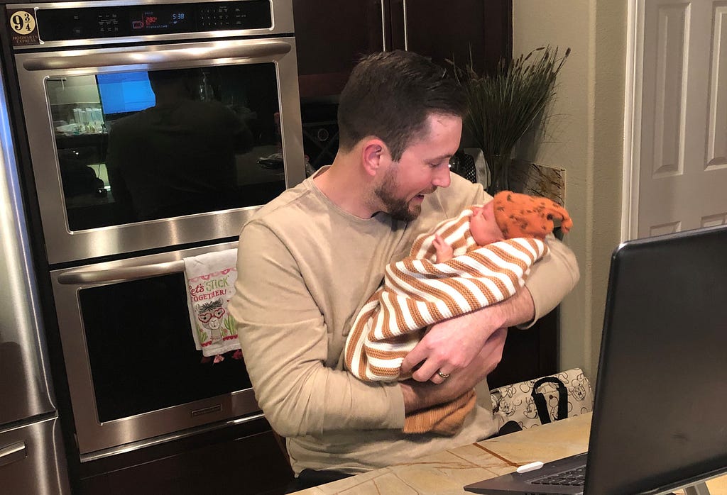 Brett holding baby Ezra in the kitchen while working on a laptop. Brett is looking down and smiling at Ezra. A Platform 9 and 3/4 magnet can be spotted in the background.