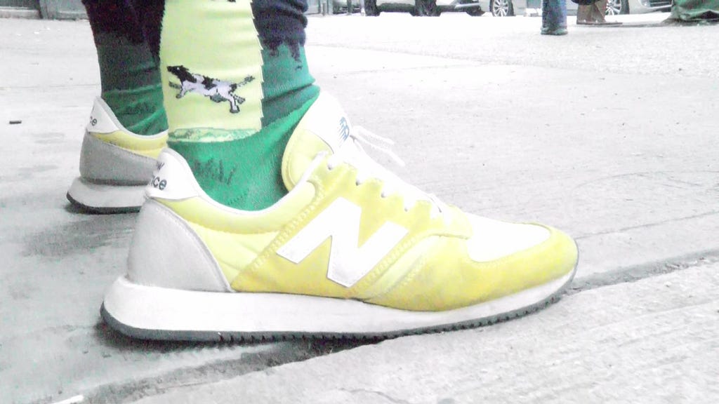 A man wearing a pair of yellow New Balance trainers with his pants rolled up to show off his socks. The socks have a design of Cows and UFOs on a green background.