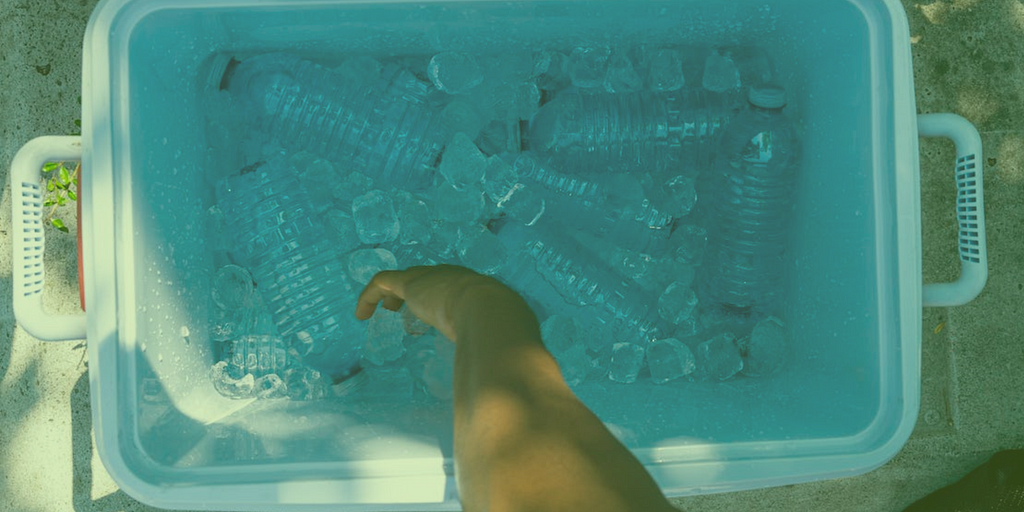 An individual’s arm reaching for a plastic water bottle in a cooler of ice and plastic water bottles.