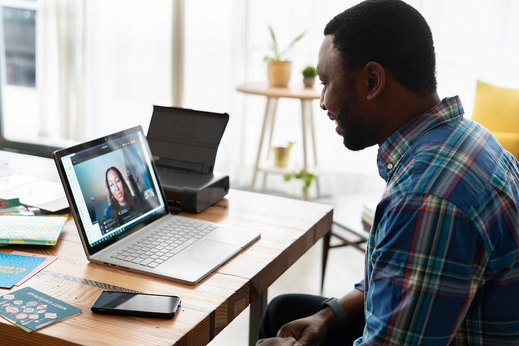 A black man staring into a laptop screen. The laptop is on a wooden desk. A woman’s face is on the laptop screen. Their posture and set-up models a virtual interview.