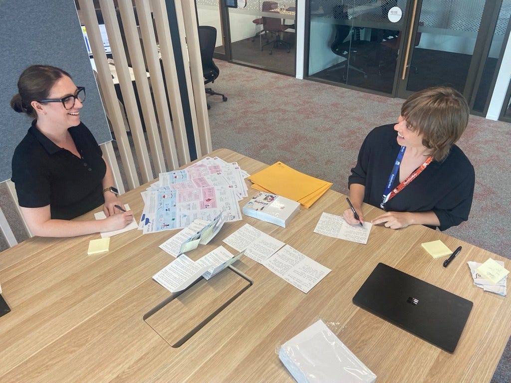 A photo of two women at a table, writing on cards. The table has a spread of cards, posters and envelopes.