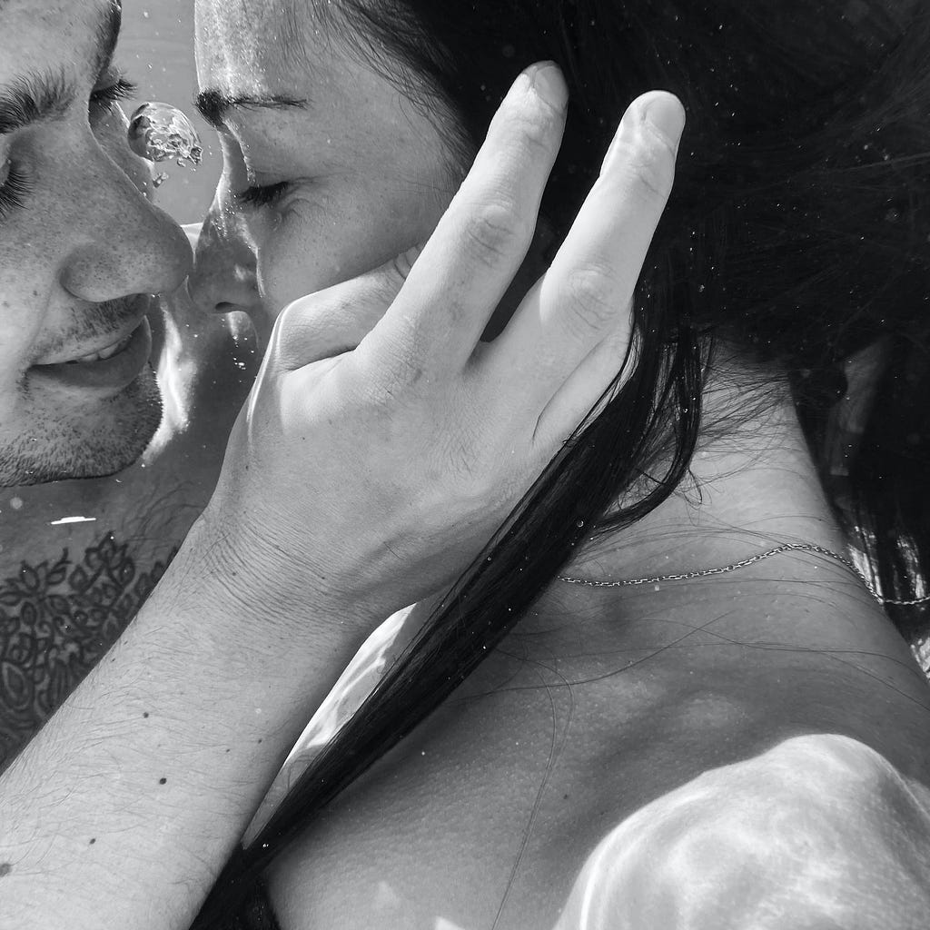 Black and white photo of two lovers with their lips almost kissing. His hand is in the forefront of the photo, gently cupping her cheek, his fingers trailing into her dark hair. They look happy and in love. She wears a thin, metal chain necklace. Her naked shoulder is visible at the bottom of the photo, and he has a patterned tattoo on his chest, barely visible in the photo. A bubble between them signifies that they’re underwater.