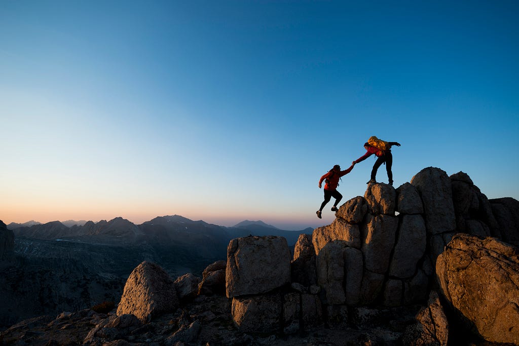 one person helping another on a climb at the top of rocks with the sun low on the horizon