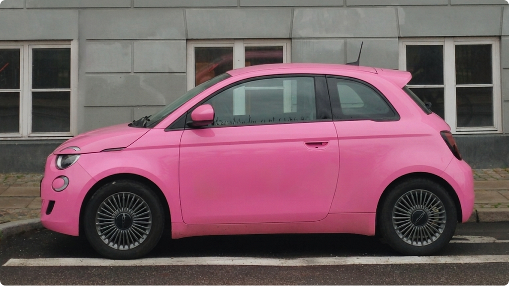 A bright pink Fiat 500 parked alongside a grey concrete building, photographed from the side in flat daylight.