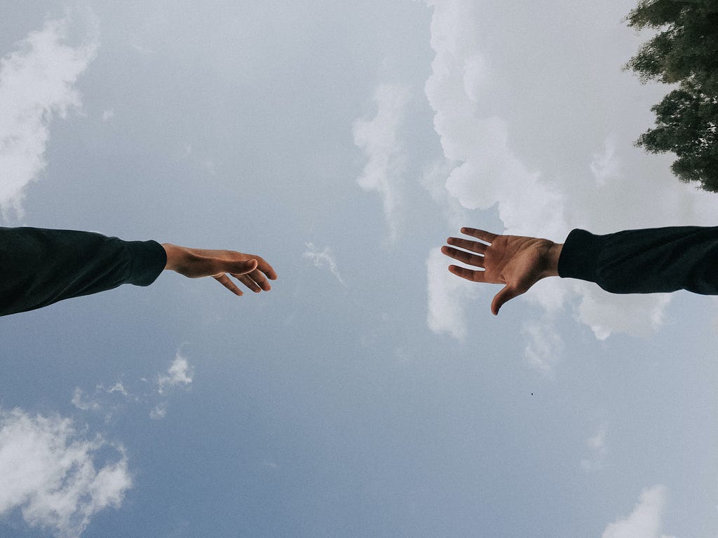 A photograph of two hands reaching for each other, with the sky in the background