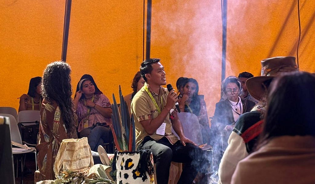 A person in a large tent is holding a mic and talking to a group of other people who are sat around listening. There is also a plume of smoke.