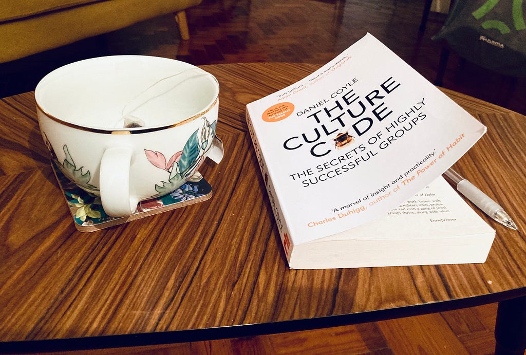 A dark wood coffee table. On the left, a white flowery tea mug, to the right a copy of the book “The Culture Code”