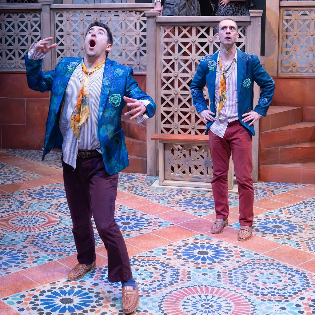 Two men dressed identically in blue jackets and red-brown pants look up; the man in front looks like he’s just realized something.