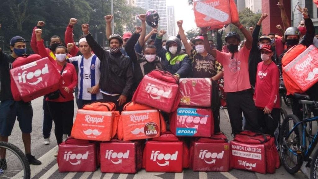 Photograph of around 20 Anti-Fascist Deliverers, all with their right fists in the air, standing behind a pile of delivery backpacks showing off the company logos Rappi and iFood at Avenida Paulista in São Paulo, Brazil.