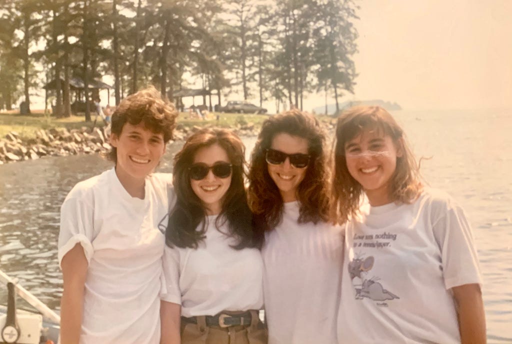 Color photo weathered from age showing 4 young brunette women arm-in-arm, posing with with bigs and a lake in the background with some land and tall pine trees, in daylight, all wearing white t-shirts, including the story author and the actress Shannen Dohert in the middle, who are both donning black sunglasses.