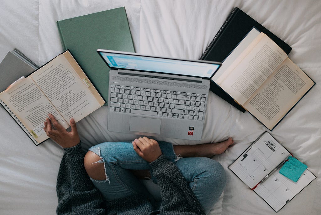 uma mulher sentada de calça jeans em cima da cama com um notebook na frente e livros ao redor.