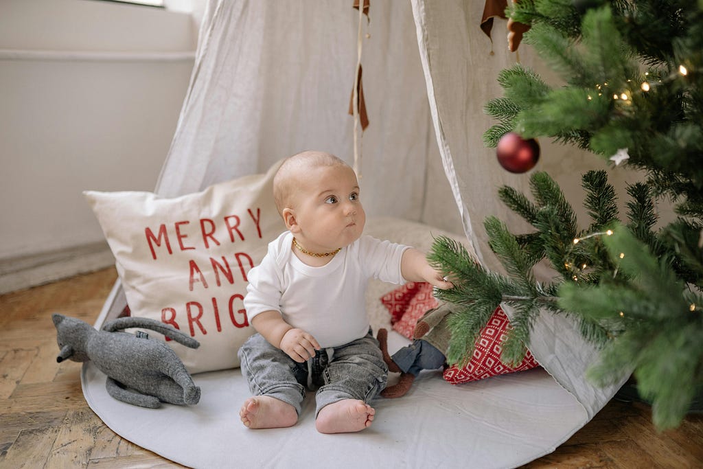 This image features a baby sitting on a soft, white surface inside what appears to be a cozy indoor play tent. The tent is made of light fabric and is decorated with hanging elements like leaves or feathers. The baby, who is looking up to the left with a curious expression, is wearing a simple white long-sleeve top, grey pants, and has a golden necklace.