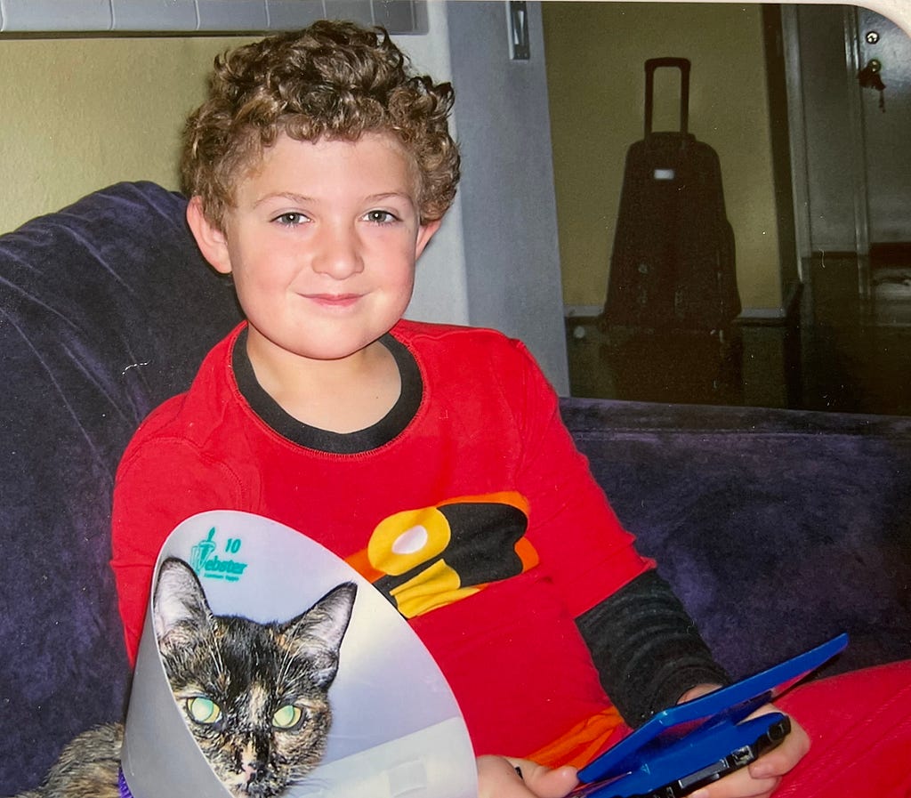 A picture of me at 8 years old, sitting on a couch with my cat playing my original, blue Nintendo DS. I am smiling at the camera.